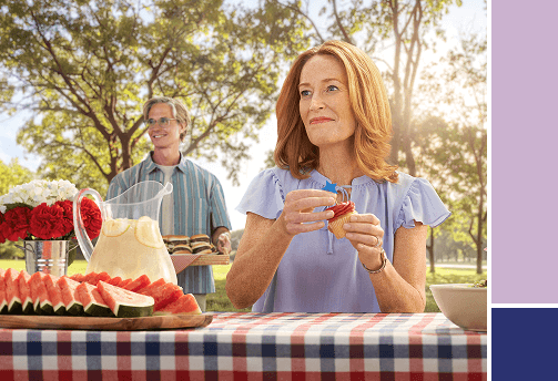 A woman at a picnic table puts a blue star on a cupcake, with watermelon and lemonade on the table, while a man brings a tray of food in the background.