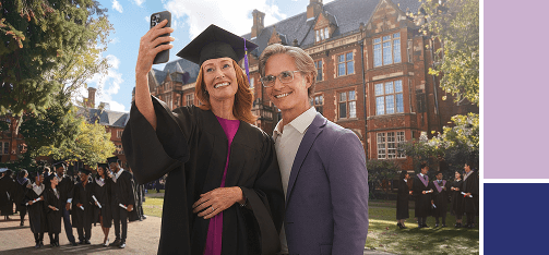 A smiling woman in a black graduation cap and gown takes a selfie with a man on a university campus. In the background, there is a large brick building and other graduates celebrating.