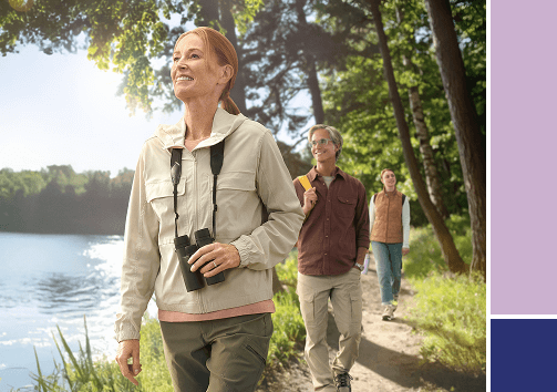 A woman with binoculars smiles while hiking on a sunny trail next to a lake with two other people.