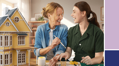Two women painting a yellow dollhouse.