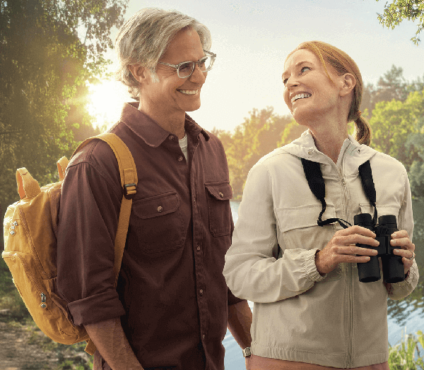 A man with a yellow backpack and a woman holding binoculars smile at each other while hiking near a lake.