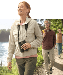 A woman with binoculars smiles while hiking on a trail next to a lake with two other people.