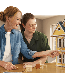 Two women smile while furnishing a yellow dollhouse together at a table, one of them placing a miniature chair inside.